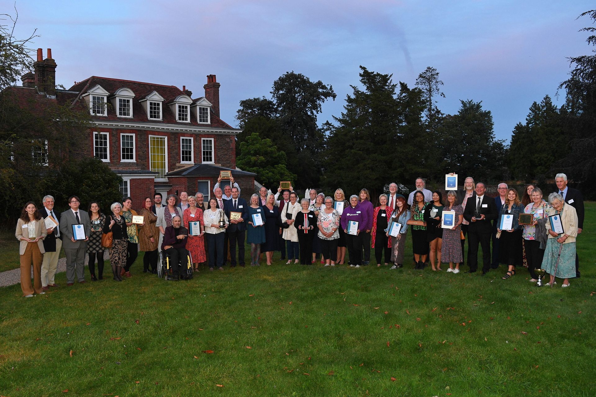 A large group of people holding awards and certificates stands on a lawn in front of a historic building at dusk.